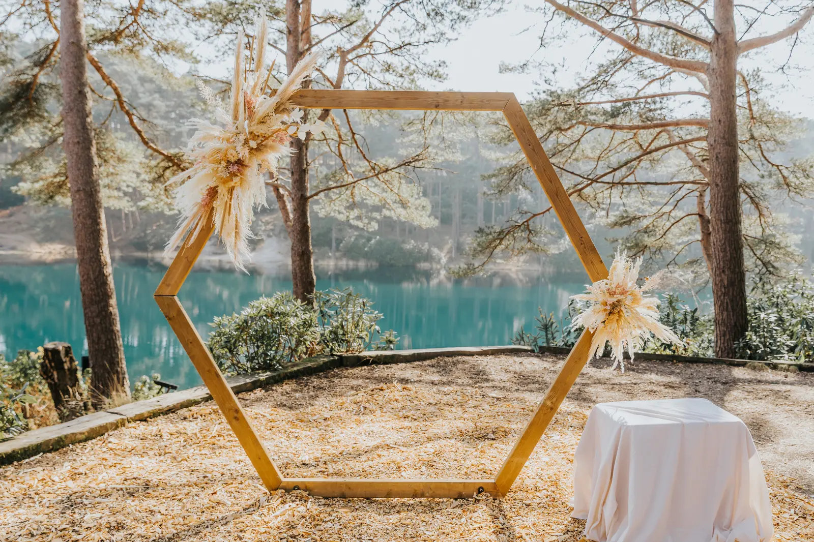 A wooden hexagonal arch dressed with pampas grass stands on a sunlit clearing beside the turquoise water of The Blue Pool, with log benches set out in front.