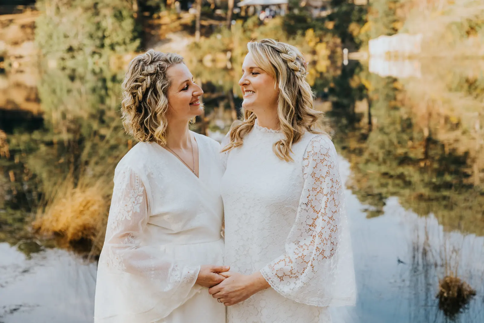 Two brides in lace dresses laugh together on the shore of the lake, pine trees reflected in the water behind them.