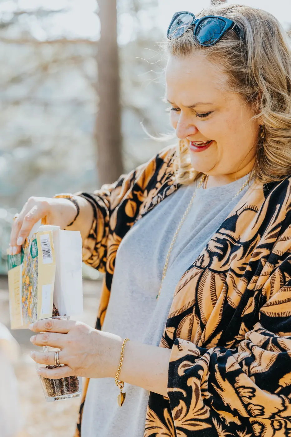 A celebrant in a patterned jacket smiles as she pours sand into a glass vessel during a blending ceremony.