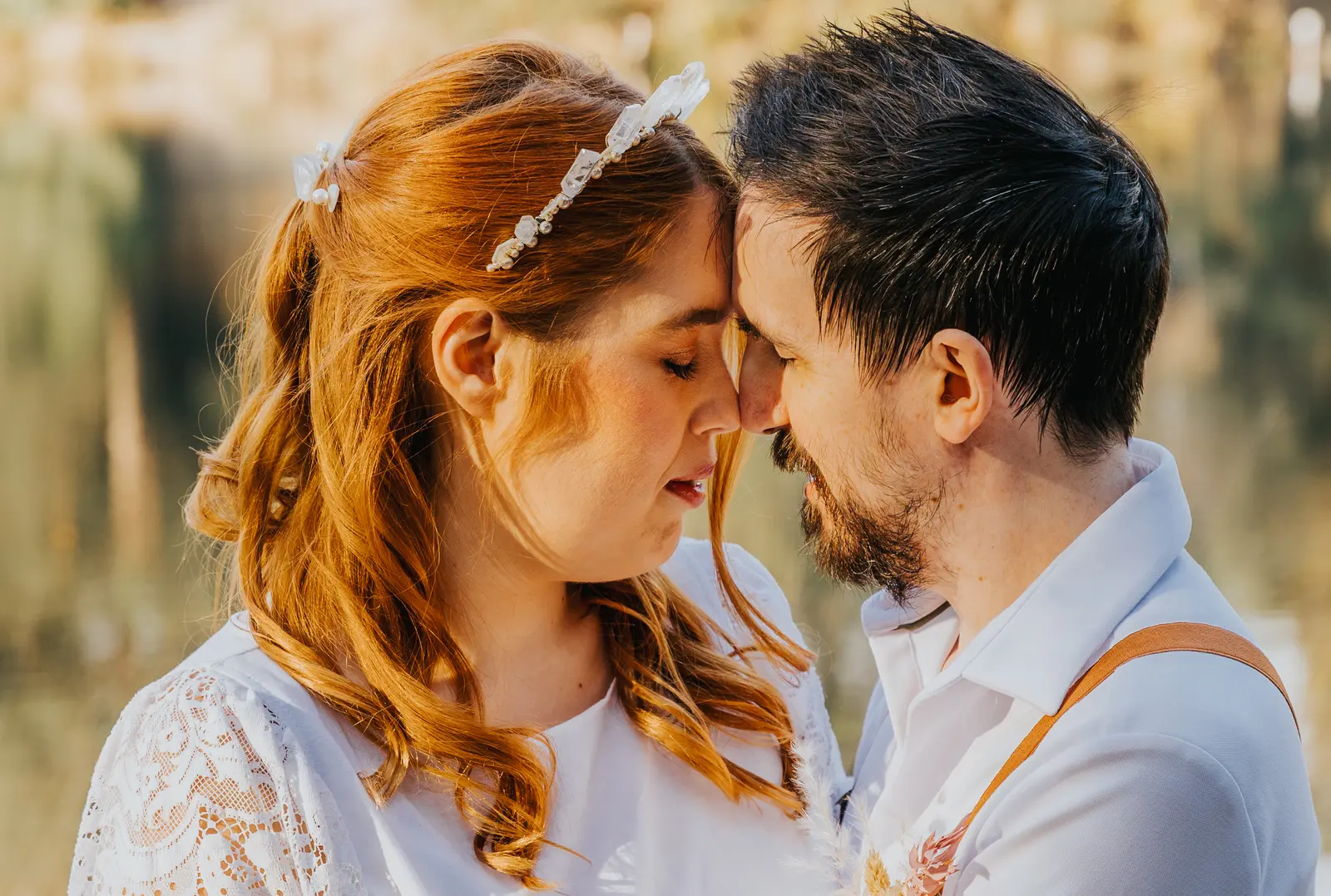 A couple rest their foreheads together, her red hair pinned with small white flowers, his braces over a white shirt.