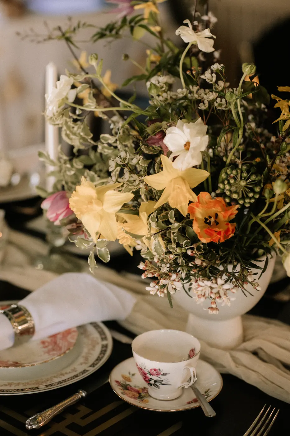A loose arrangement of garden flowers in a white jug on a laid table, with vintage china and a candle.