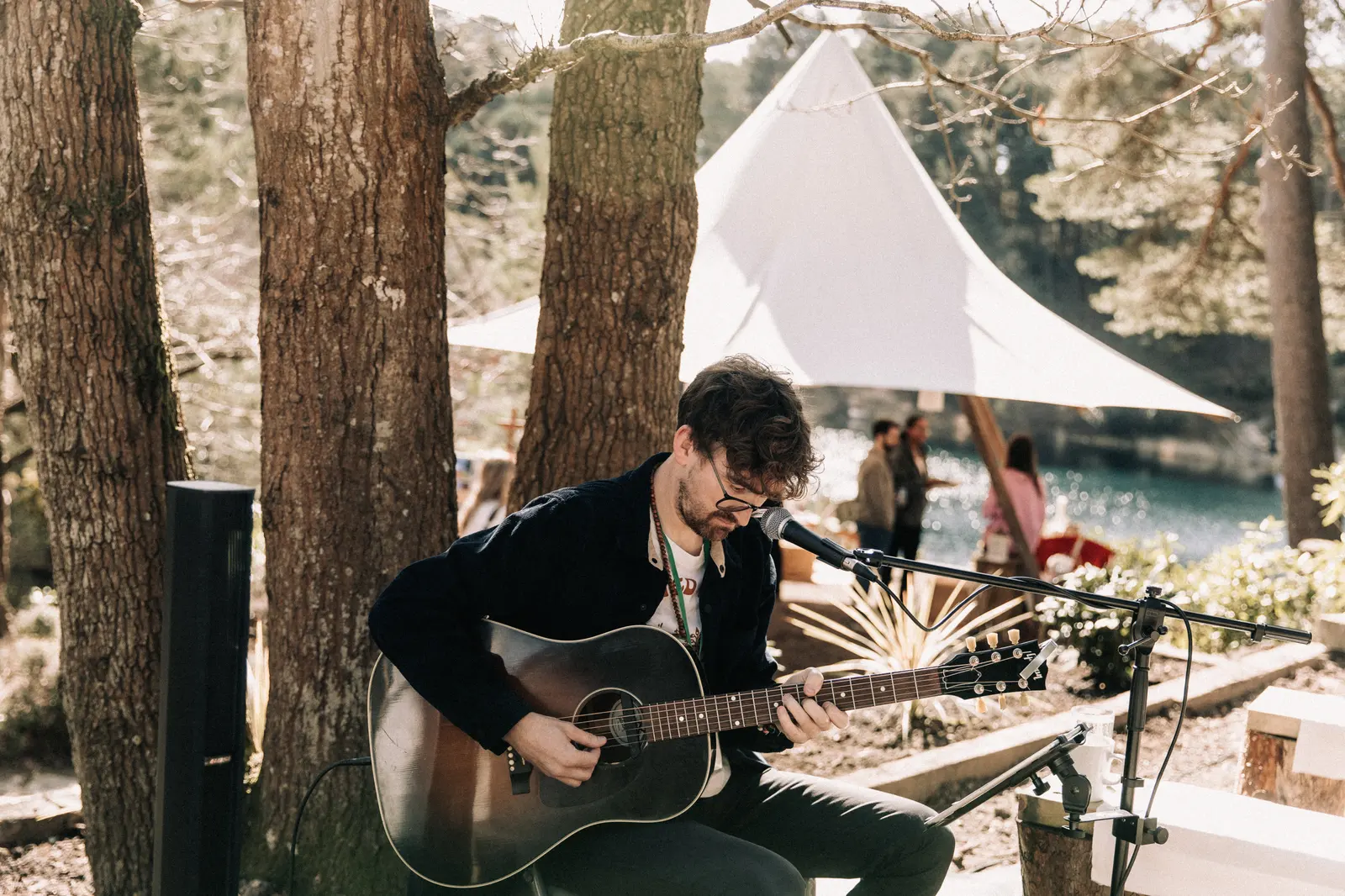 A musician plays acoustic guitar beneath the pines, a white tipi behind him.