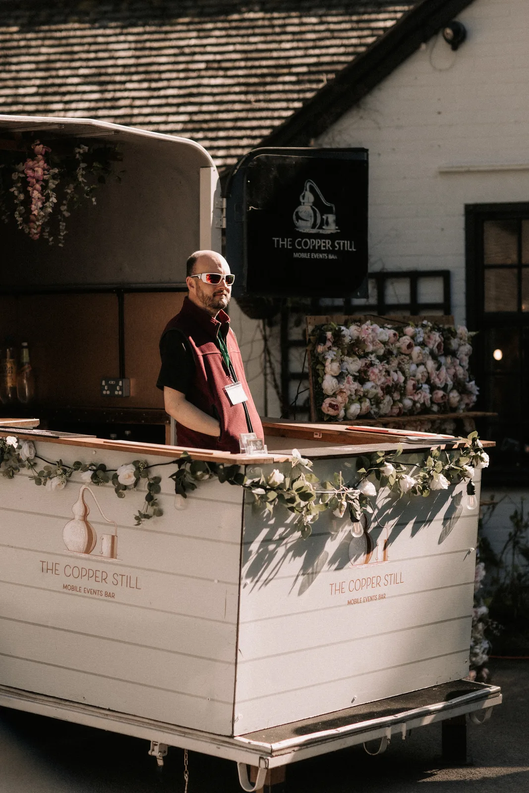 A white mobile bar dressed with foliage and festoon lights, the bartender behind it.