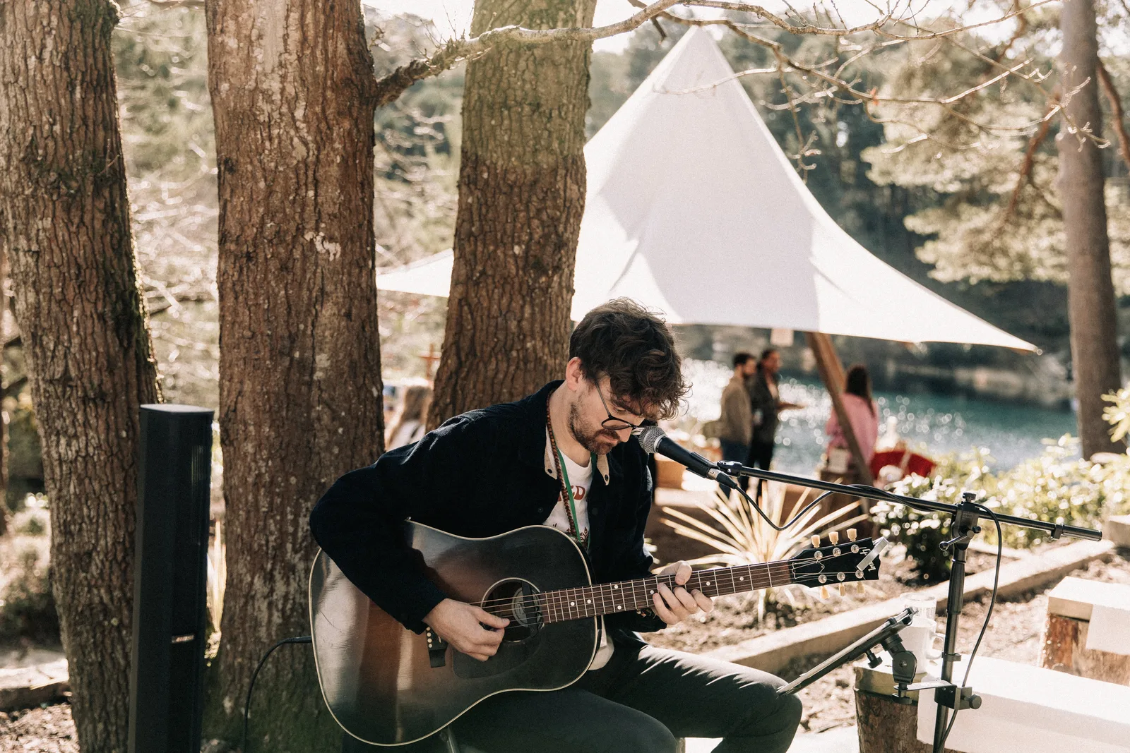 Leigh Coleman plays acoustic guitar under the pines, a white tipi and the lake behind him.