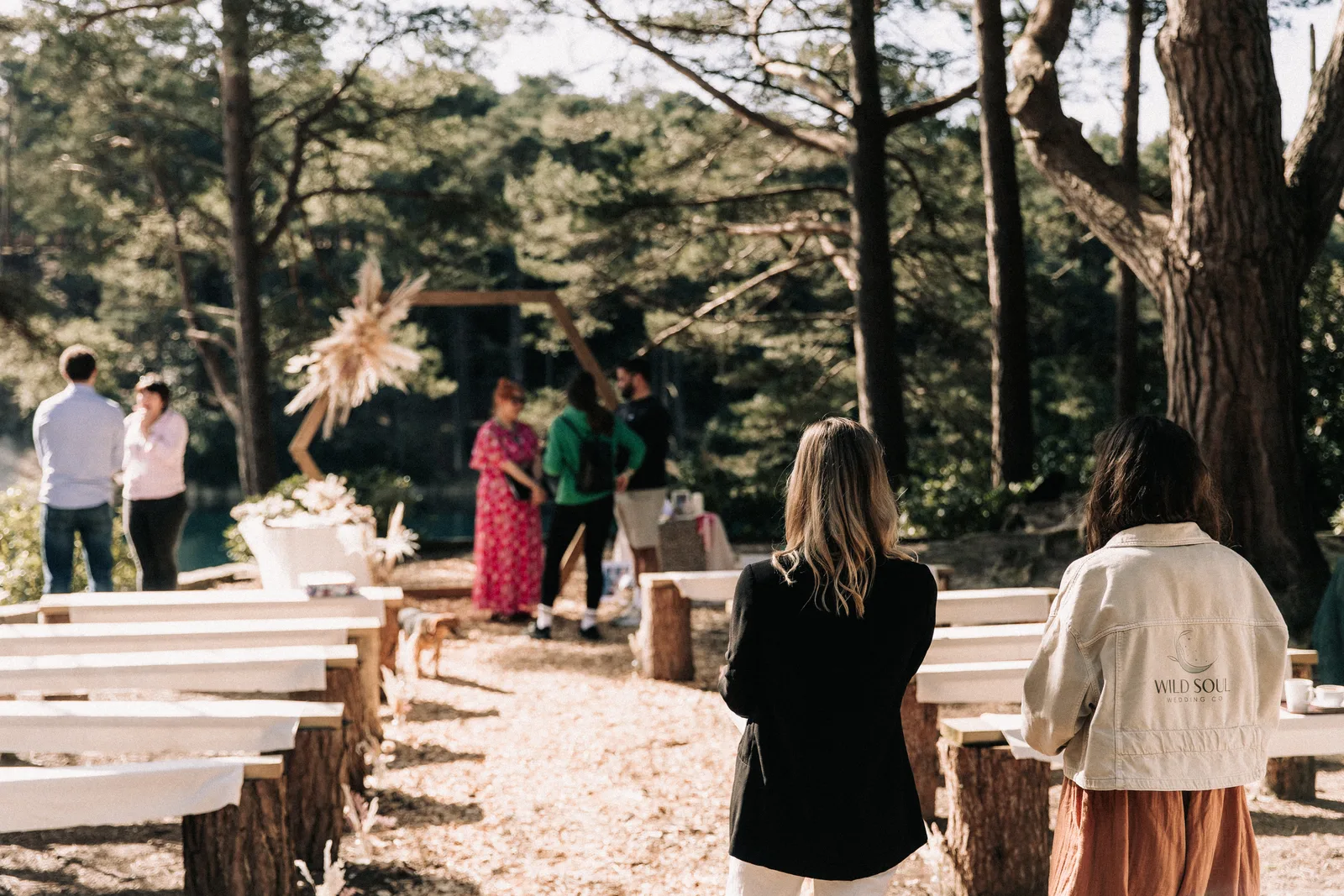 Couples wander between log benches in the ceremony clearing, a dried-flower arch at the far end.